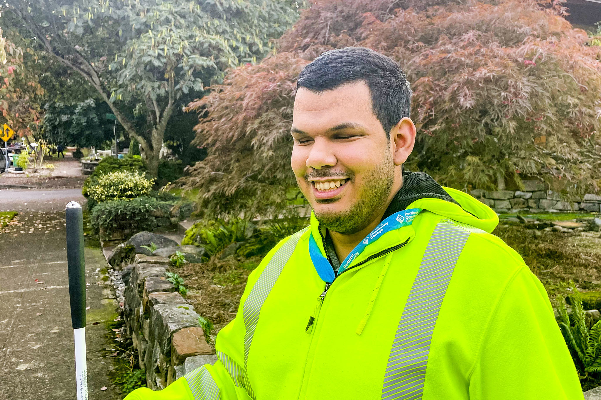 A portrait photo of a smiling latino man from the shoulders up, wearing a reflective jacket outside in front of some bushes.