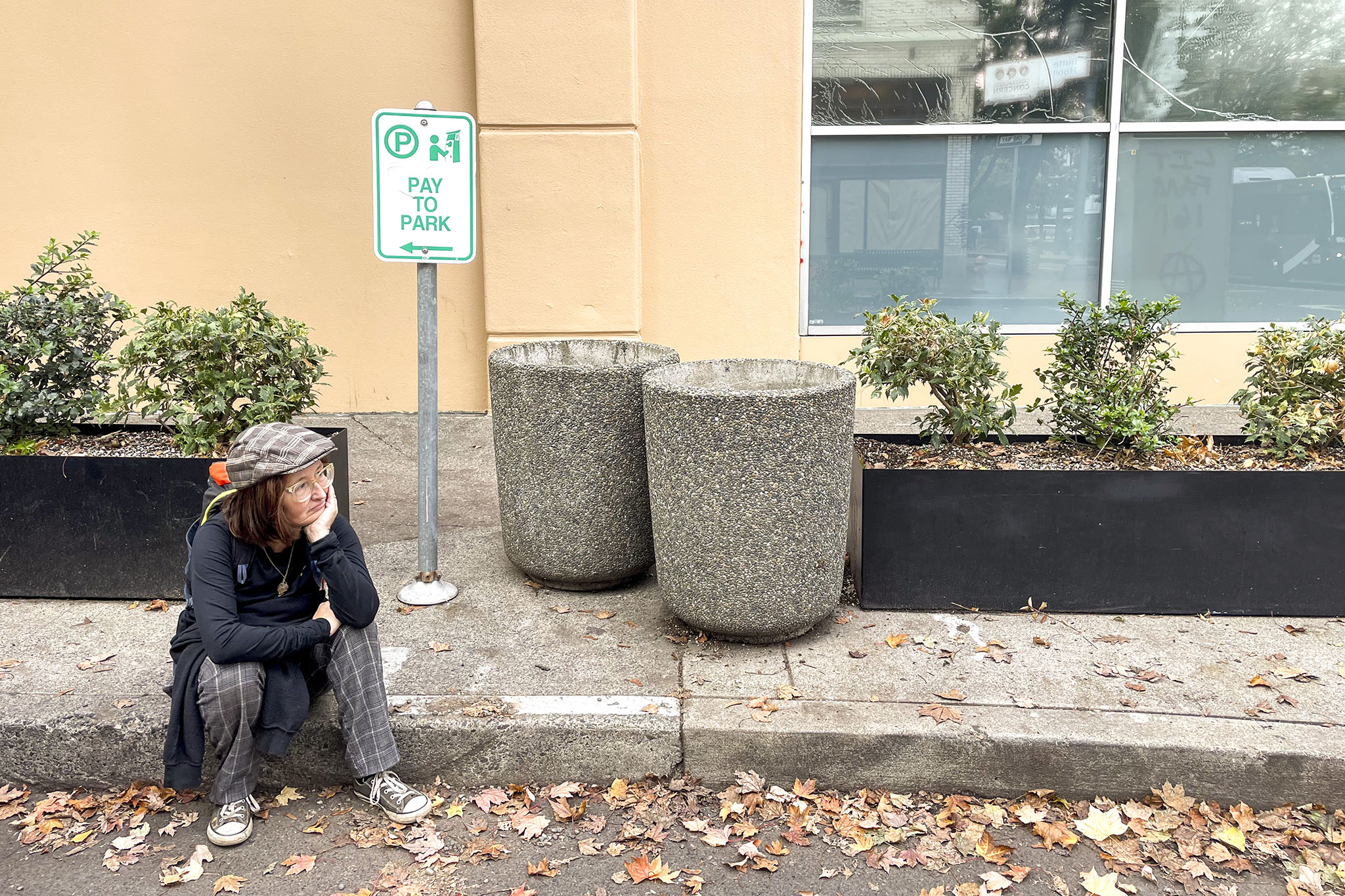 Barbra Weber sits on a sidewalk in downtown Portland across the street from the former Sisters of the Road cafe, next to a “Pay to Park” sign and a couple of planter boxes. 