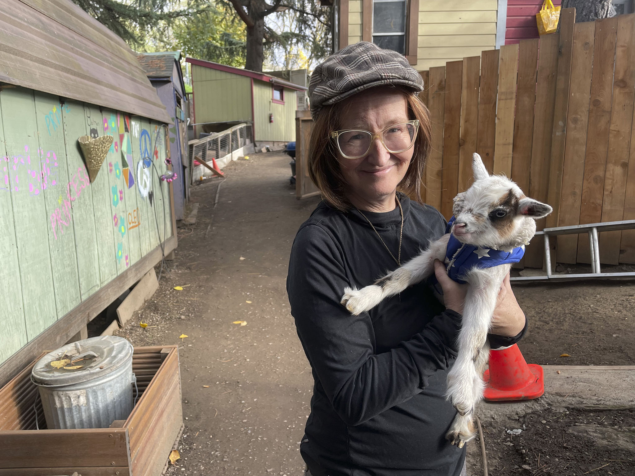 Barbra Weber holds a goat wearing a blue zip-up jacket for dogs. The rest of the neighborhood is visible in the background. 