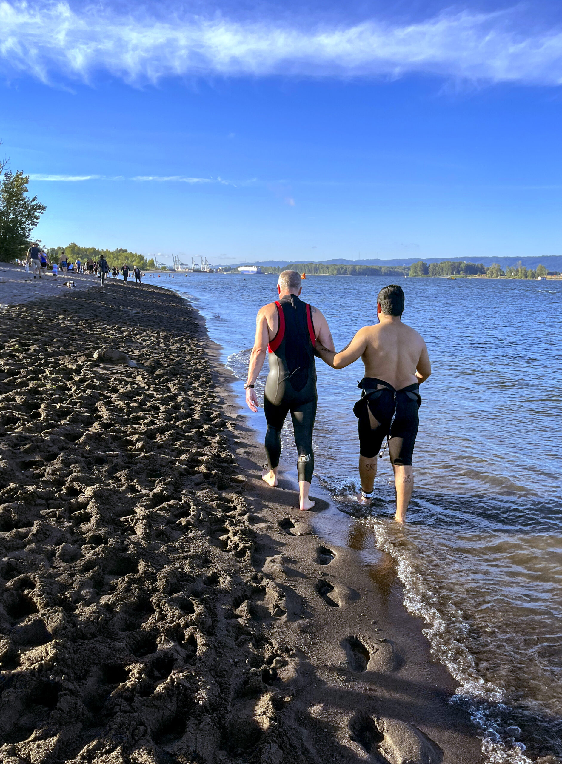 Two men walk arm-in-arm along a beach, both wearing wetsuits. Footprints in the sand trail behind them.