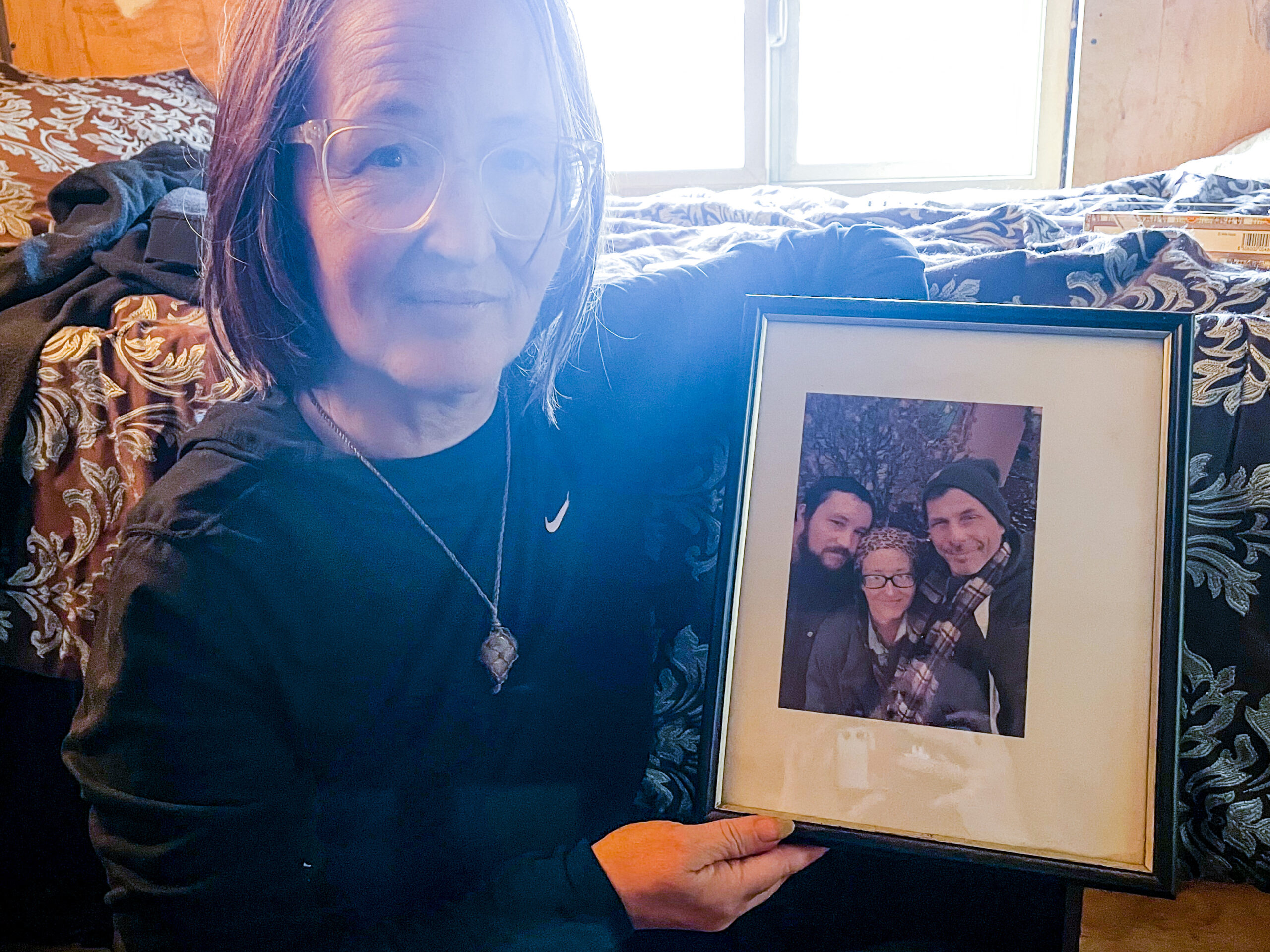 Barbra Weber stands next to her bed holding a framed photograph of herself and her son James, who died from a fentanyl and methamphetamine overdose in 2022.