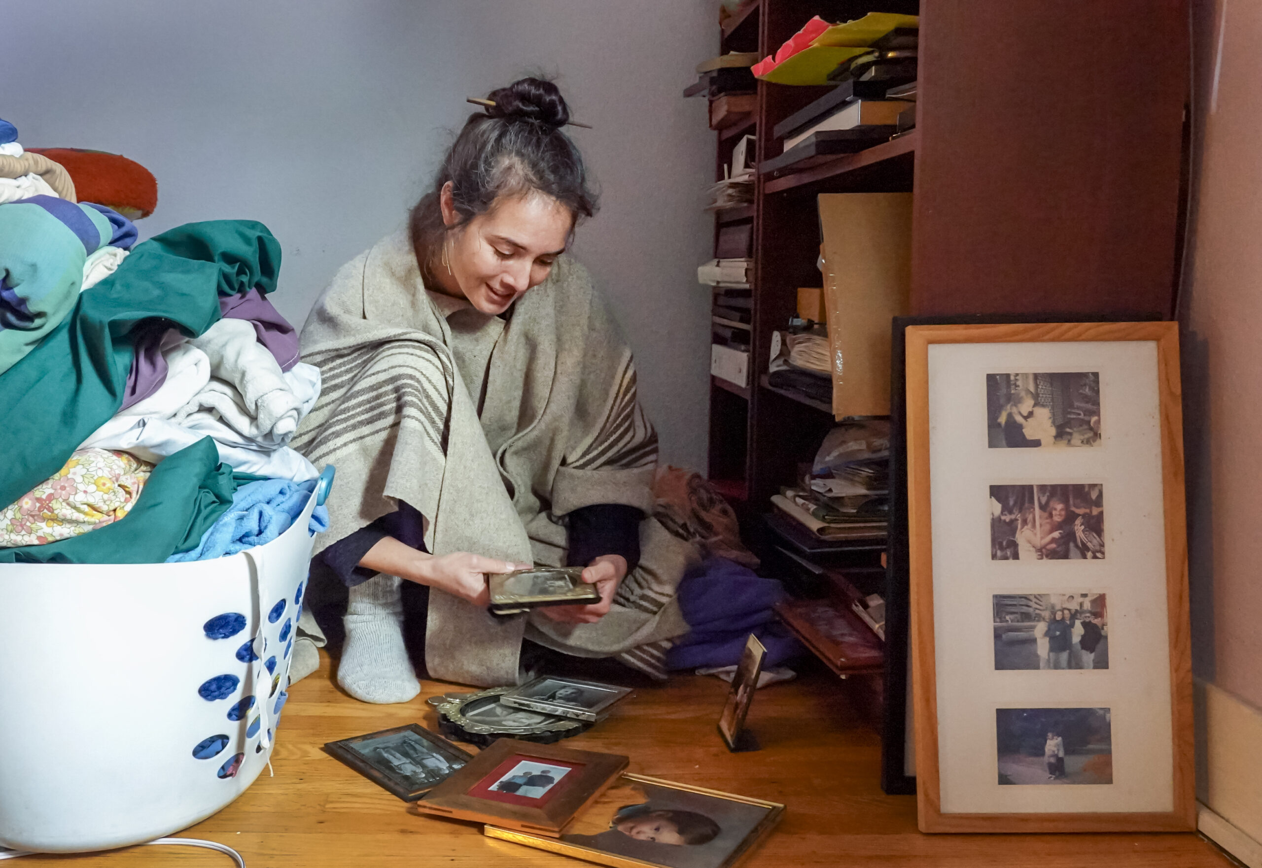 Green sits on a hardwood floor between the foot of a bed and a shelving unit, with a pile of framed photos in front of her.