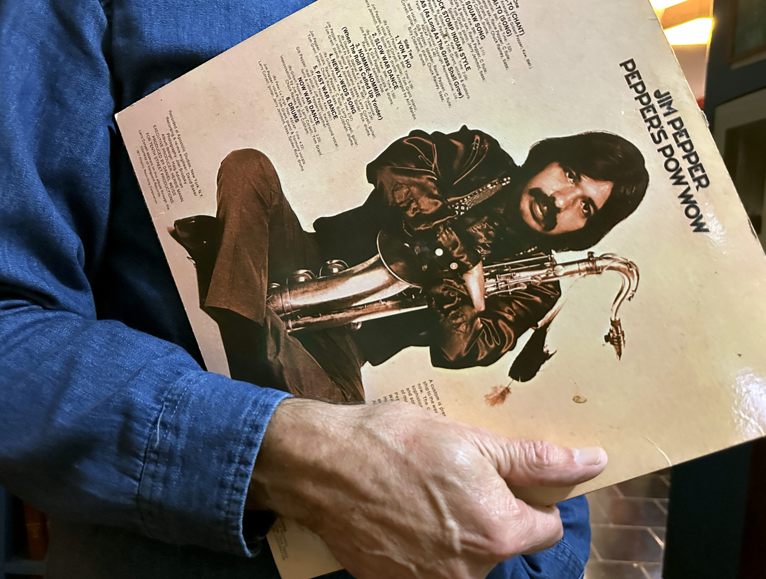 Sean Cruz holds Jim Pepper’s 1971 debut album, Jim Pepper’s POW WOW in his arms. Jim Pepper is pictured sitting cross-legged with a saxophone in his arms.