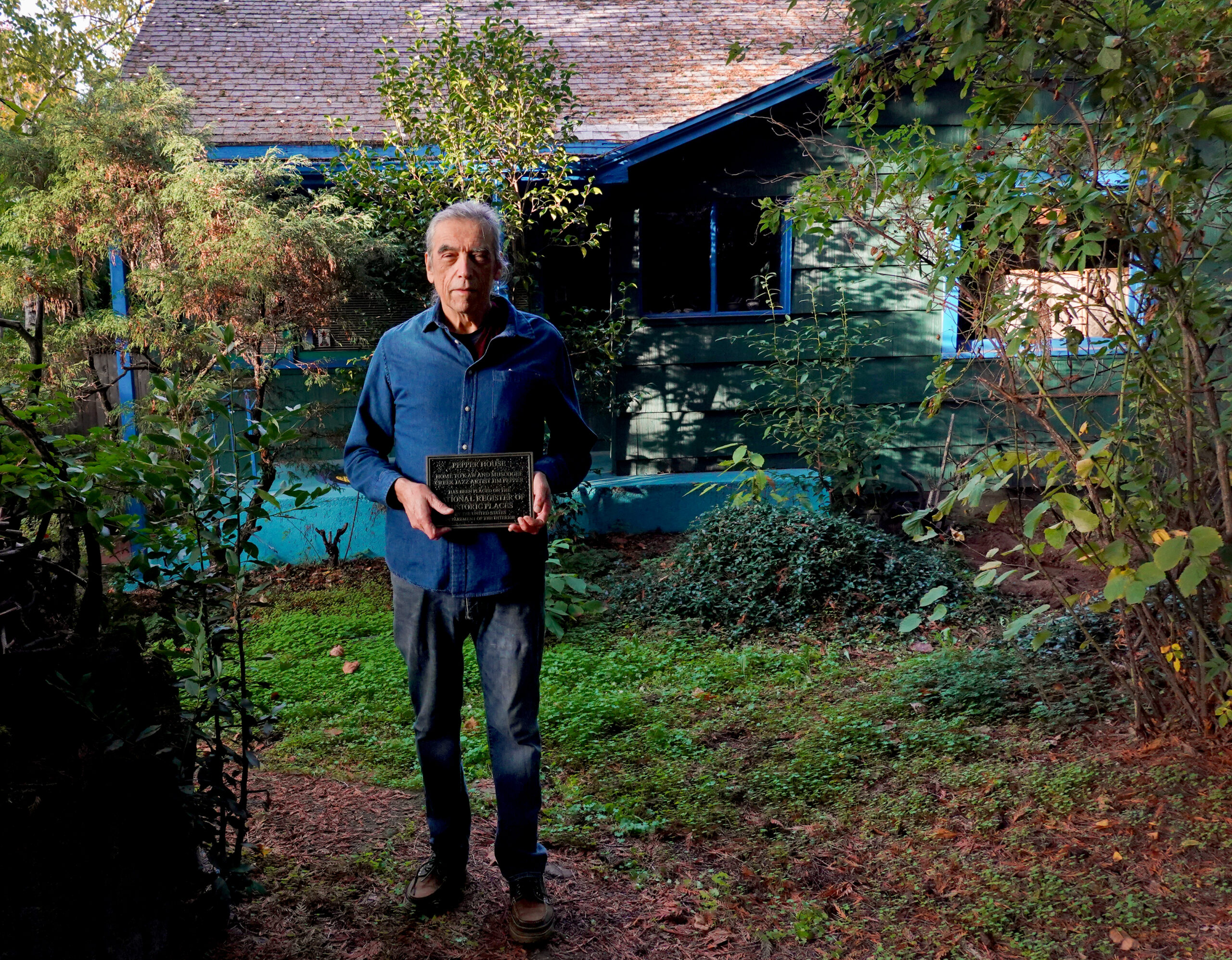 Sean Cruz in front of his house holding the plaque he received from the Department of Interior. The house is different shades of blue and green. Shrubs and trees surround the house.